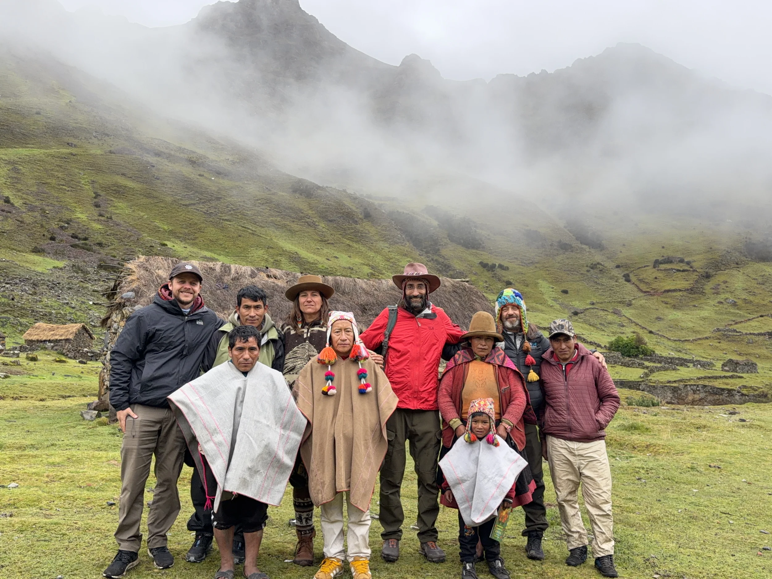 Paititi team standing with Q&rsquo;ero families outside a thatched stone house, mist coming down the Andean valley behind them