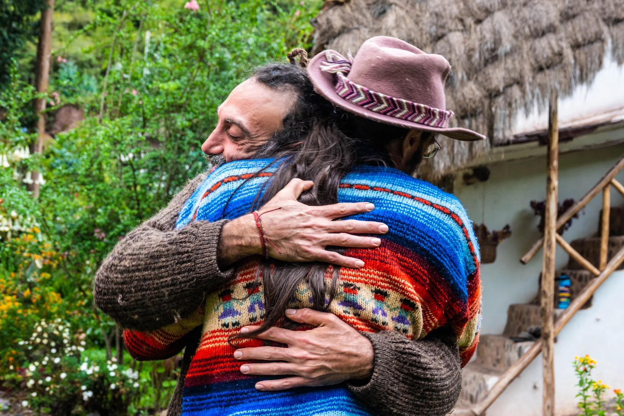 Two people hugging outdoors, one wearing a colorful striped sweater and a hat, with green foliage and a rustic building in the background.