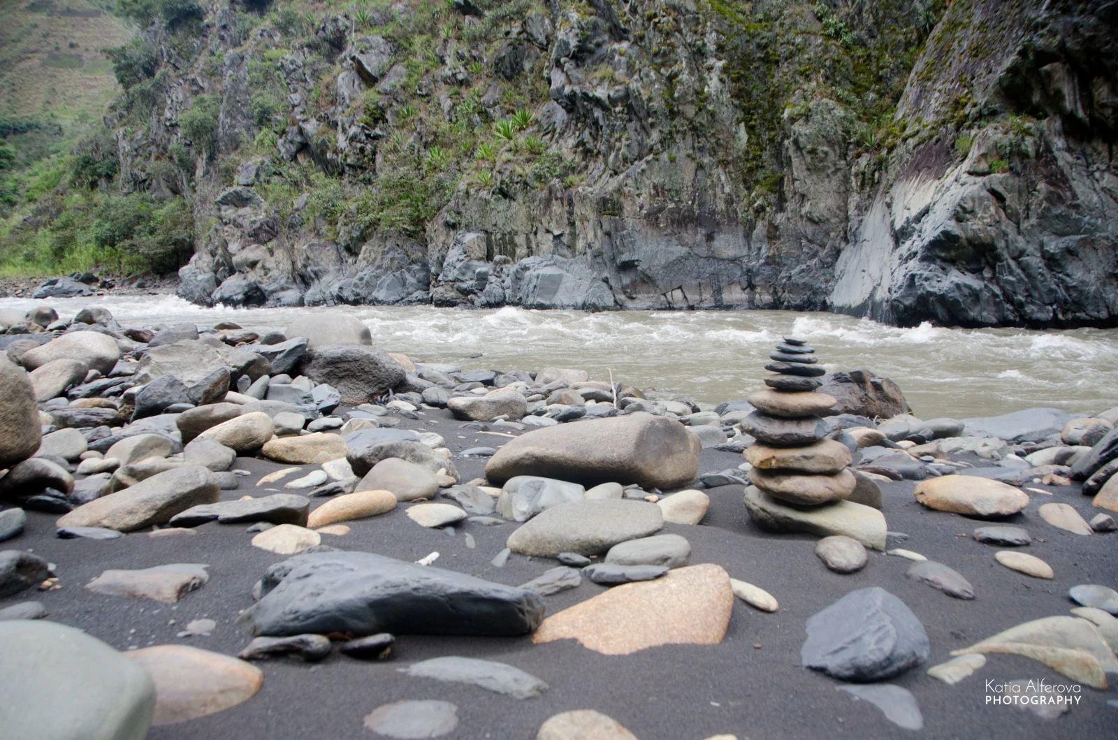 Una apacheta de piedras equilibradas sobre la arena negra de la orilla del río Mapacho, con el agua verde y rápida y los farallones detrás